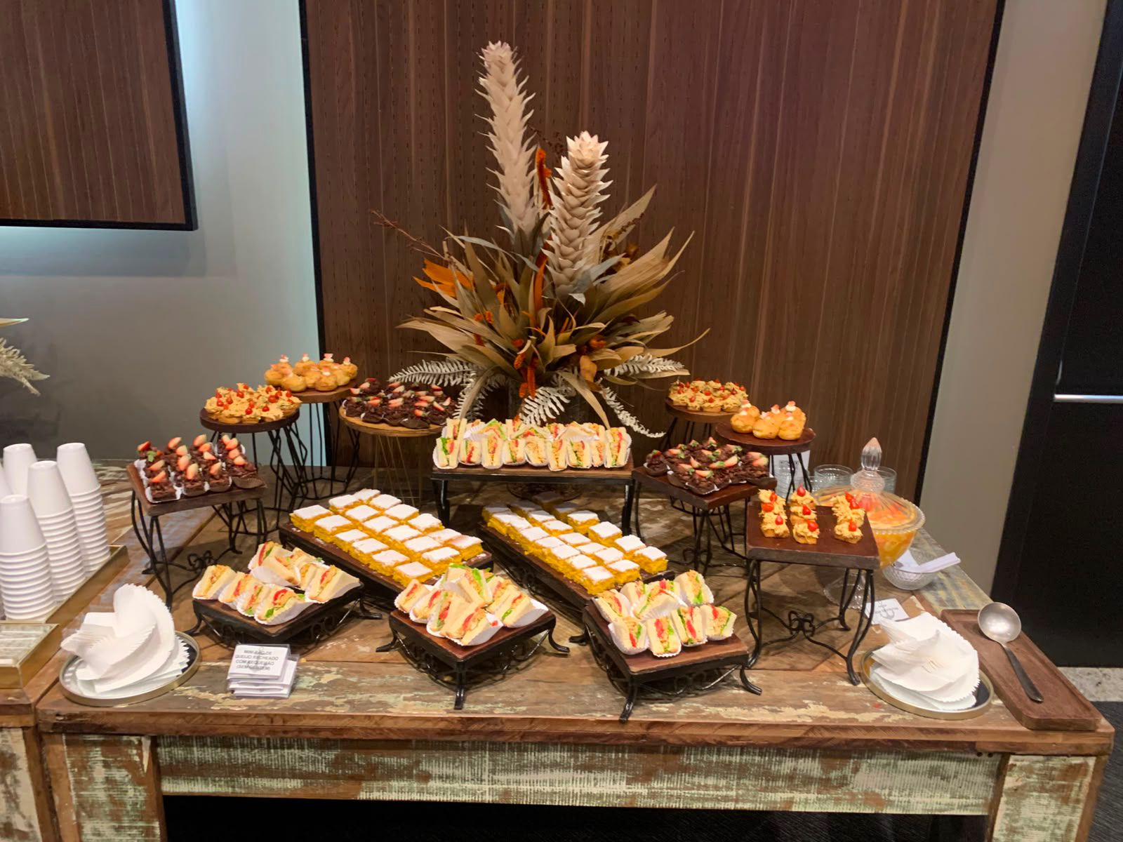 Mesa de coffee break montada com sanduíches, doces e arranjo floral seco